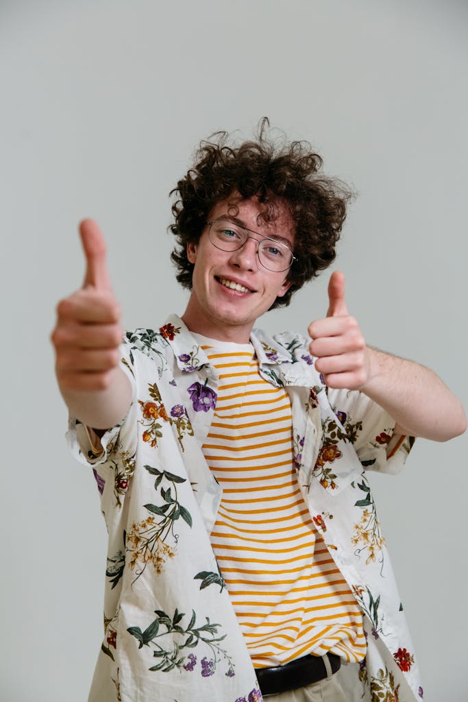 Smiling young man in floral shirt with thumbs up in front of gray background.
